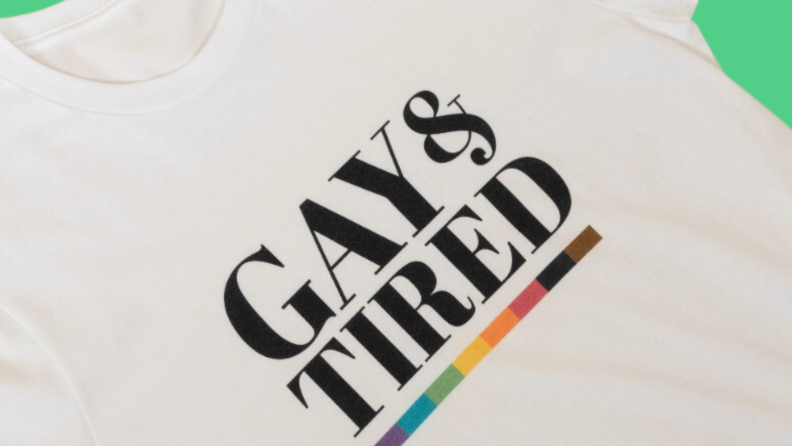 A close-up of an off-white T-shirt from Gay Pride Apparel with a strip of rainbow beneath a slogan reading, Gay & Tired.