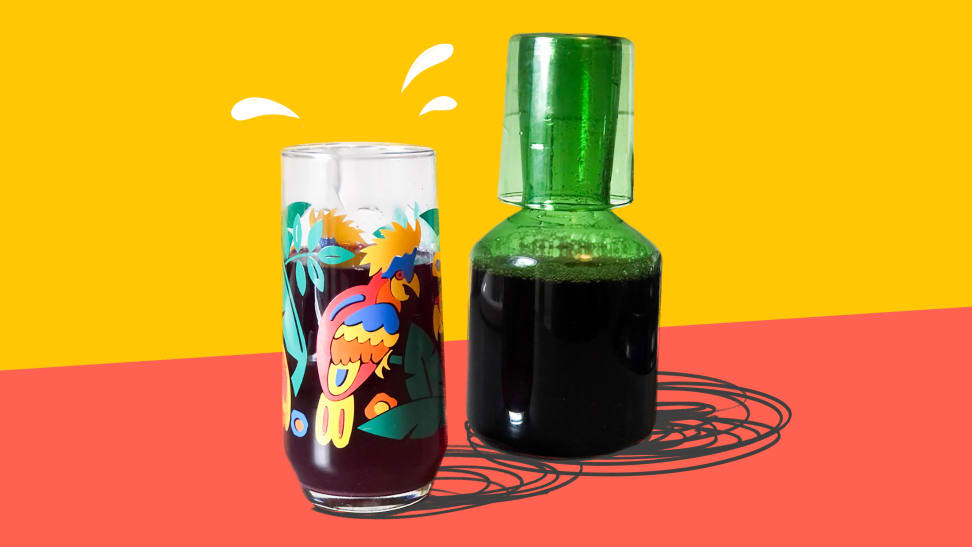 A colorful glass filled with iced hibiscus tea next to a green glass carafe filled with more of the same.