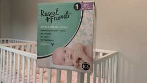 A box of Rascal and Friends size one diapers sits on the edge of a crib in a gray room