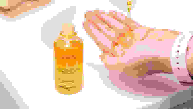 A bottle of yellow-colored hair oil sitting on a sink and a hand with oil on it next to it.