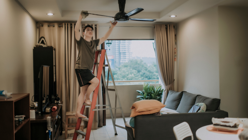 A person standing on a ladder cleaning a ceiling fan.
