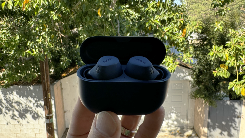 A hand holding the blue Jabra Elite 8 Active earbuds in their open case with an alley and trees in the background.