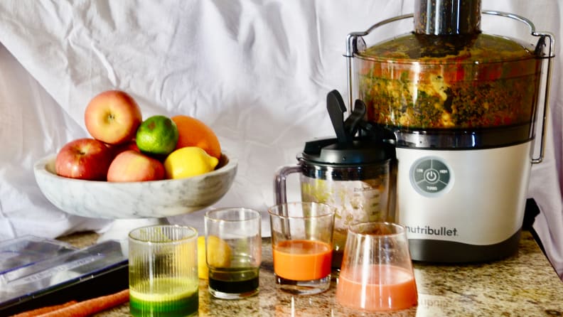 The NutriBullet Juicer Pro sits on a counter in a kitchen and is full of produce being juiced. Four glasses of different colored juices and a bowl of produce sit in the foreground.