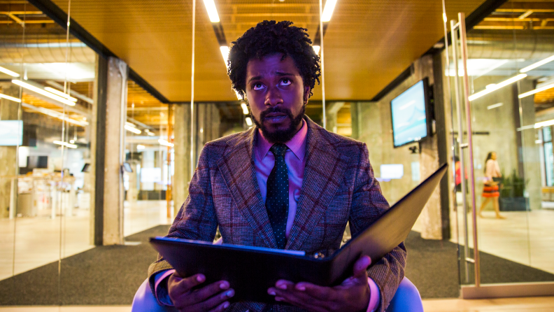 Actor LaKeith Stanfield sits—holding a three-ring binder and wearing a suit—in the 2018 dark comedy Sorry to Bother You.