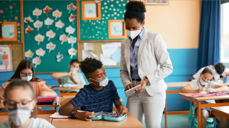 Teacher wearing face mask while helping student in face mask in classroom setting.