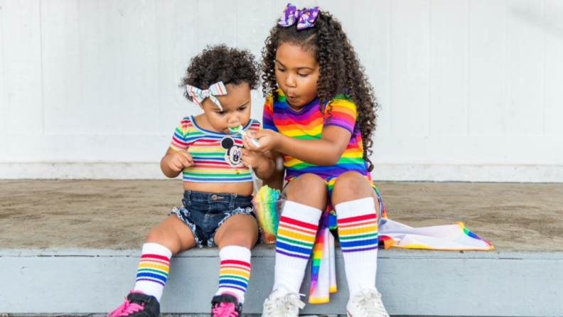 Two little girls sitting together outdoors sharing a snow cone while wearing rainbow attire.