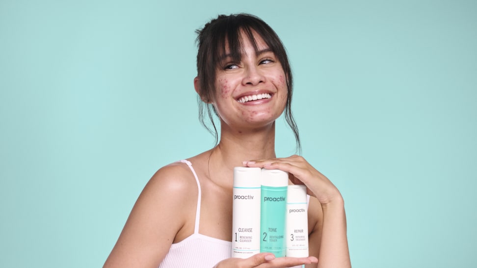 A smiling young woman with acne holds up three proactiv acne treatment products against a teal background.