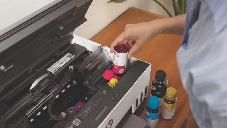 A close-up of a person refilling an HP Smart Tank printer with magenta ink, with other color ink bottles arranged on a wooden desk nearby.