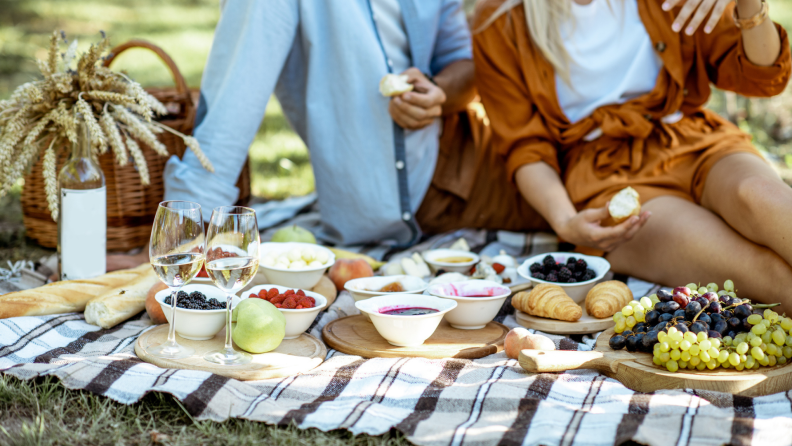 A couple enjoys an idyllic outdoor picnic, complete with grapes, bread, fresh fruit, and a bottle of wine.