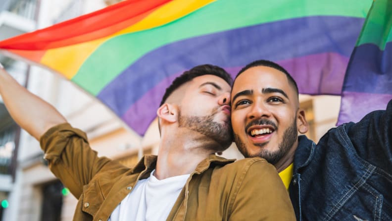 Two men holding a big rainbow flag, one kissing the other on the cheek