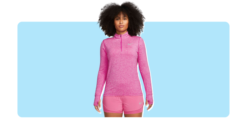 Model with curly hair wearing the long-sleeve, pink Nike Element ½-Zip Running Top with thumbholes and pink bike shorts.