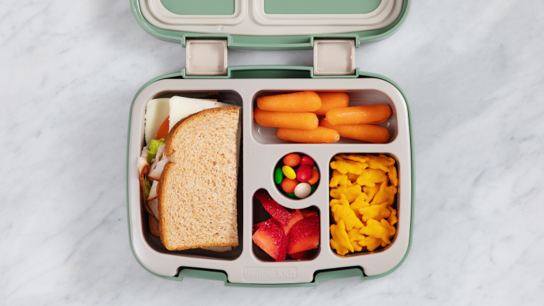 A lunch bag container with half a sandwich, fruit, veggies, Goldfish, and M&M's.