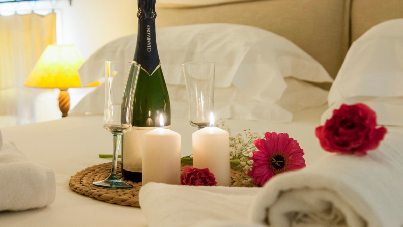 A Valentine's Day arrangement on a bed: towels, flowers, two lit candles, a pair of glasses, and a bottle of Champagne.