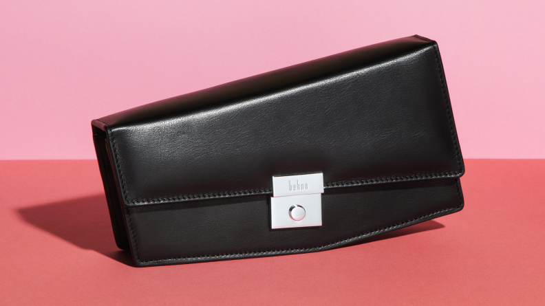 An angular black purse sits on a pink surface, the straps removed so it can serve as a clutch purse.