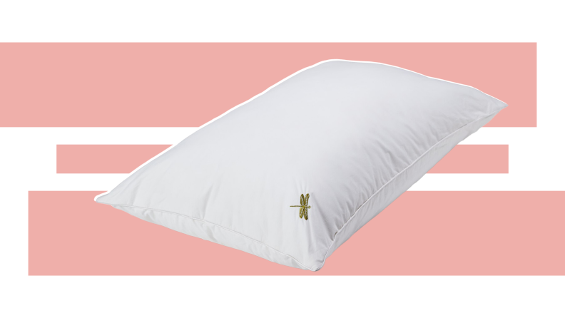 A white DreamPad pillow with a small dragonfly design embroidered in the corner in front of a pink and white background.