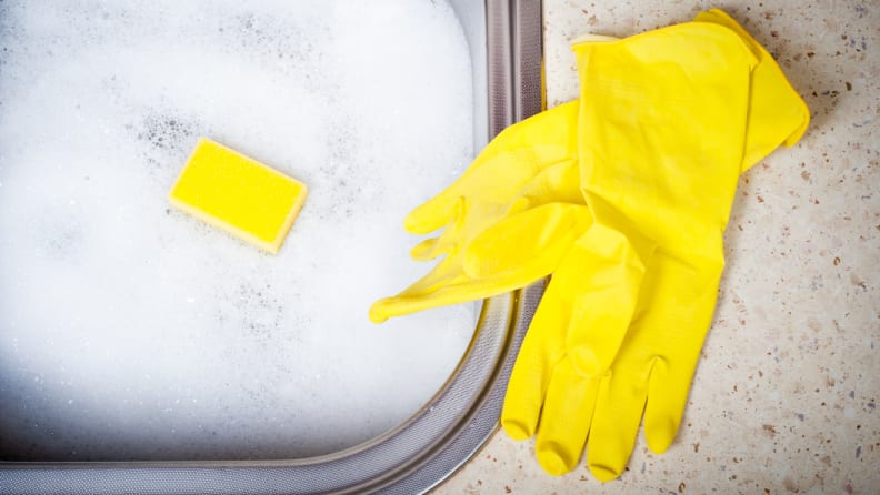 Some yellow rubber gloves sit next to a sink filled up with soapy suds. A yellow sponge is floating on top of the water, peeking out of the suds.