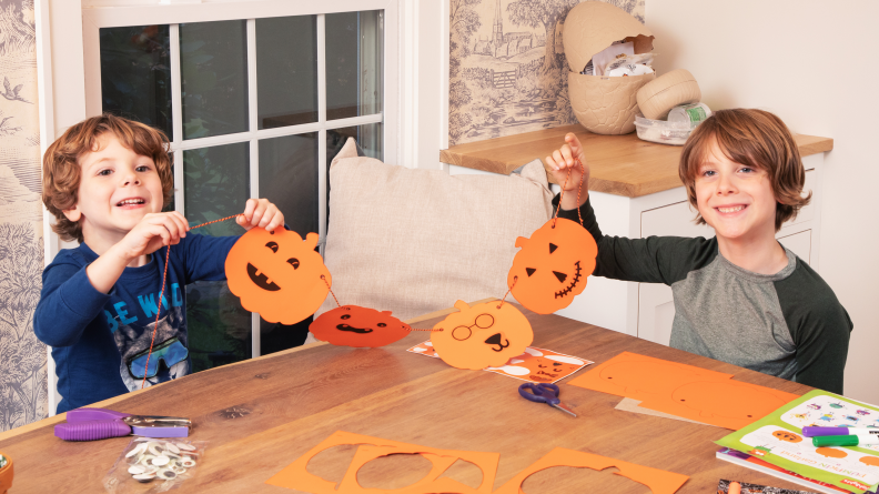 Two kids at a table holding up a pumpkin craft they made.