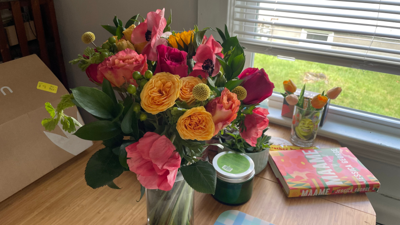 An UrbanStems bouquet on a wood table with a smaller vase of flower in the background, next to a window and book.