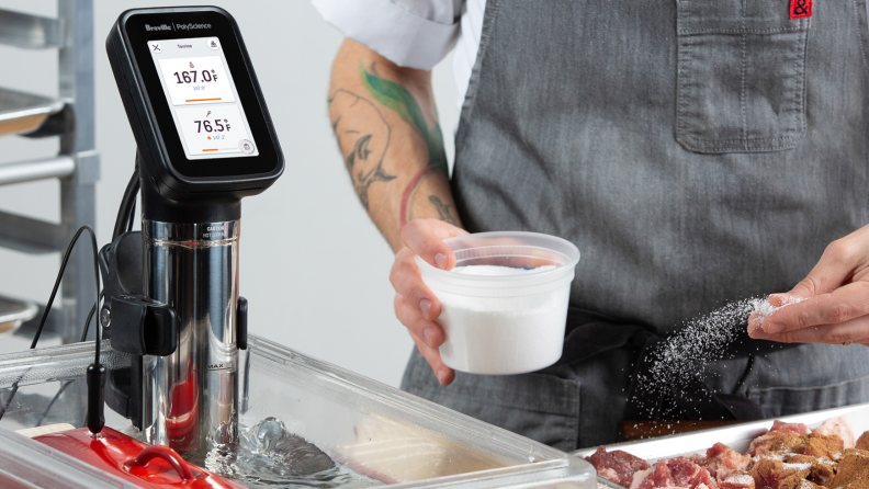 The image is centered around a PolyScience by Breville sous vide immersion circulator, which is on. An apron-wearing person is holding a container of salt, ready to season some meat.