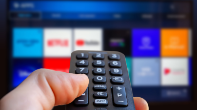 A man is holding a remote control of a smart TV in his hand. In the background you can see the television screen with streaming entertainment apps for video on demand.