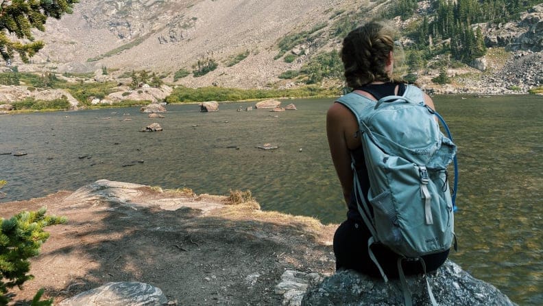A hiker stops to rest beside a body of water. Trees are scattered in the distance, on the other side of the lake.