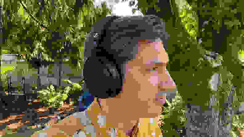 A man in a yellow pattern shirt listens to a black pair of headphones in a green yard.