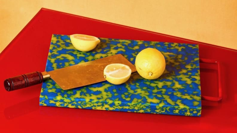 A multi-colored plastic cutting board sits on a red surface with halved lemons and a large cleaver perched on top.