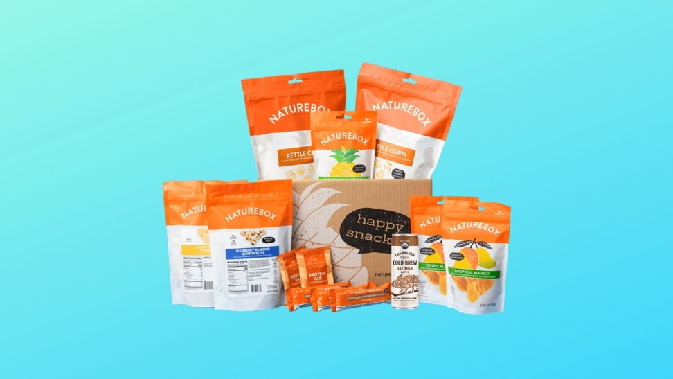 An assortment of NatureBox snacks are clustered together in front of a blue gradient background. The snacks are packaged in white bags with text on the front and a thick orange band at the top. Snacks include blueberry almond quinoa bites, dried mango, kettle corn, and more.