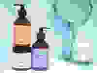 Two pump bottles a jar of hair care sit on a tile vanity next to a container of cotton swaps and a mirror.