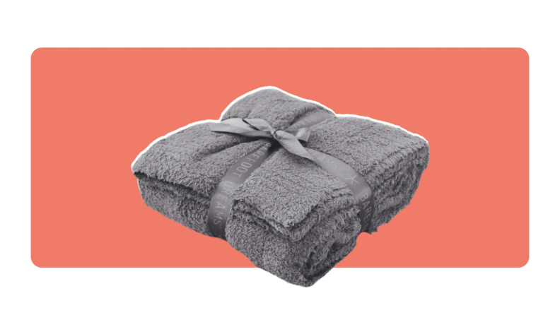 A fuzzy, gray Barefoot Dreams CozyChic throw blanket folded into a square with a gray ribbon wrapped around it.