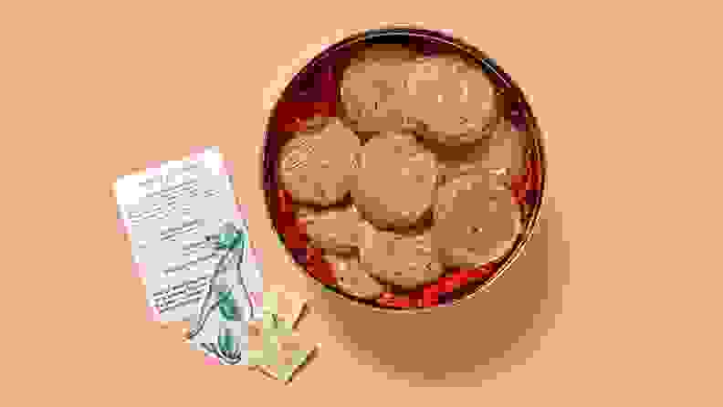 A tin of chocolate chip cookies individually wrapped in plastic wrap sits on a pink board, accompanied by matchbooks and an information sheet.