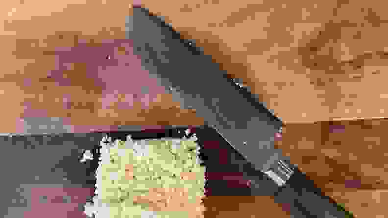 Hexclad's Damascus chef's knife with green handle next to square pile of rice on top of wooden cutting board.
