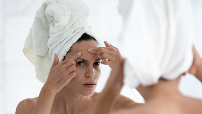 A person wearing a towel on their head, looking into a mirror, and touching their forehead.