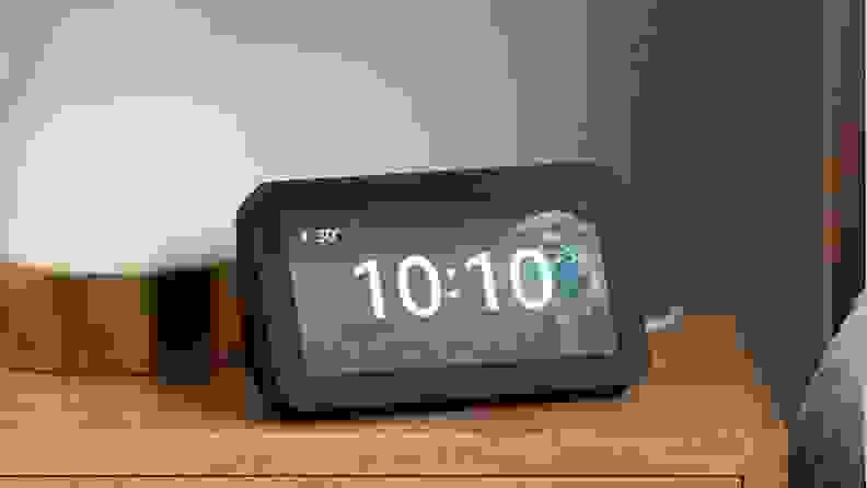 The third-gen Amazon Echo Show 5 with the time displayed on the screen, sitting on a wood nightstand.