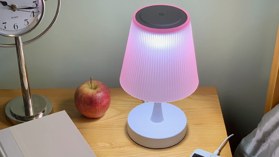 A small pink lamp illuminates a wooden nightstand beside a bed. An apple, a hardcover book, and an iPhone sit near it.