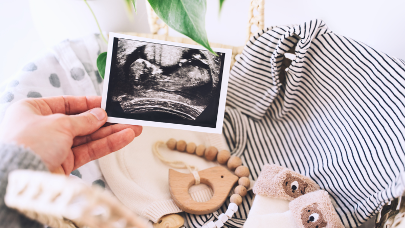 Woman's hand holding ultrasound images in background of wicker basket of stuff for newborn baby.
