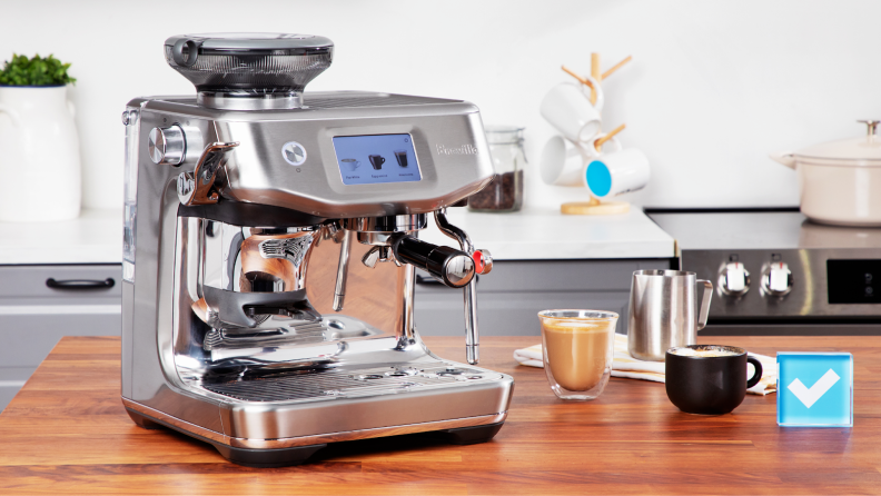 Breville Barista Touch Impress espresso machine in stainless steel finish sitting on top of wooden countertop next to two cups of coffee, an acrylic paperweight and a steel frothing pitching.