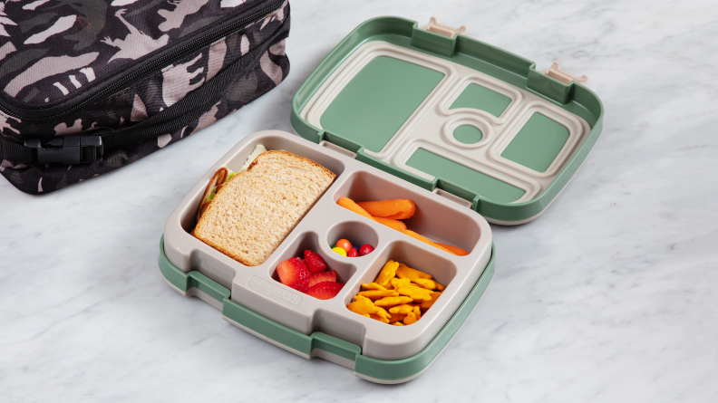 The Bentgo 5-compartment box sits open on a marble counter with half a sandwich, carrots, cheddar bunnies, strawberries and M&Ms inside. The PackIt Freezable lunch box sits in the background.