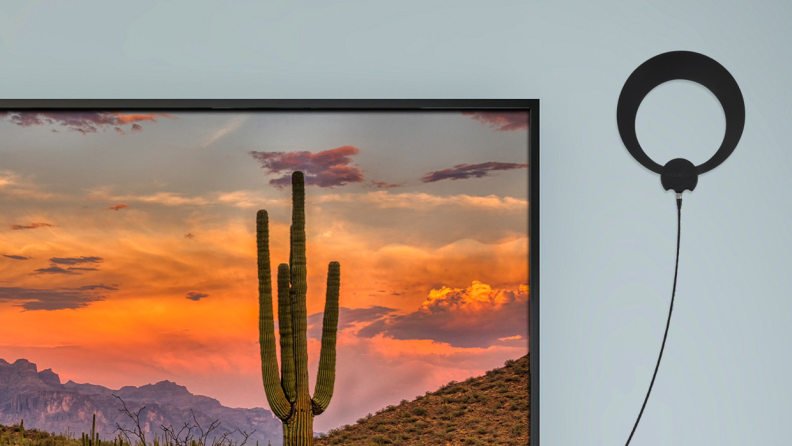 A black Antennas Direct ClearSteam Eclipse Indoor TV antenna mounted on the wall next to a TV displaying a desert landscape.