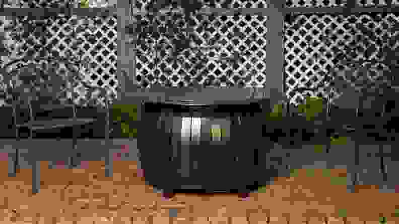 A fire-pit table is situated at the center of a brick patio. It’s flanked by two black metal chairs.