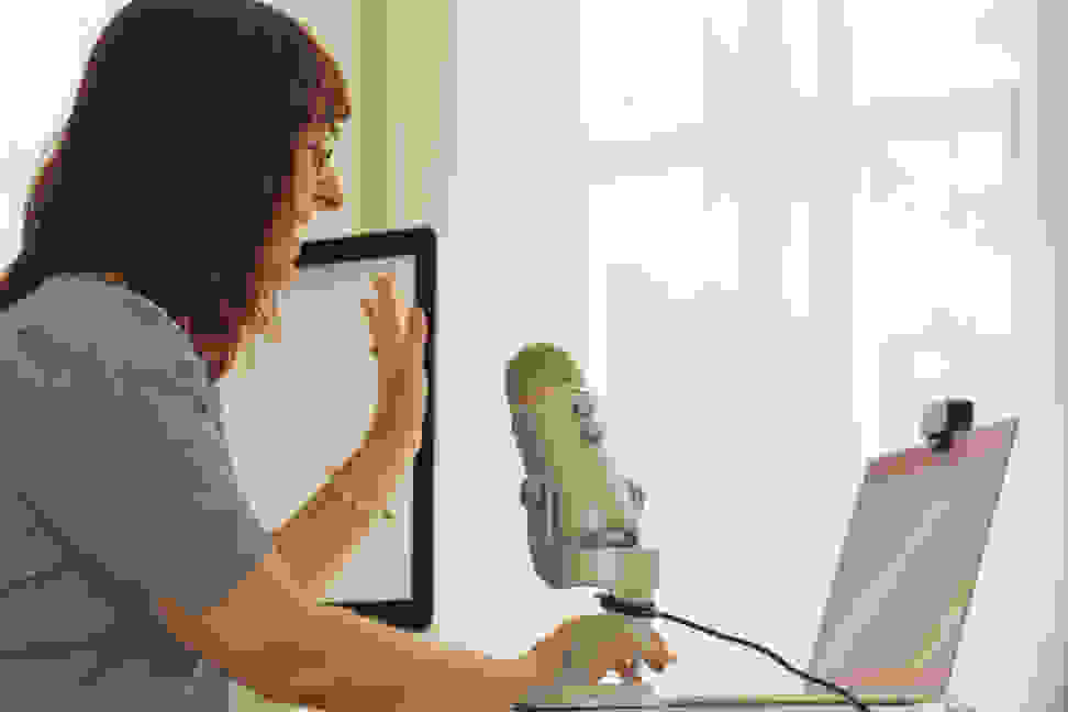 A woman waves hello at her laptop with a large USB microphone in the foreground.