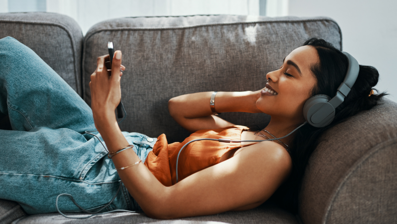 Person smiling on couch while holding smartphone and wearing headphones.