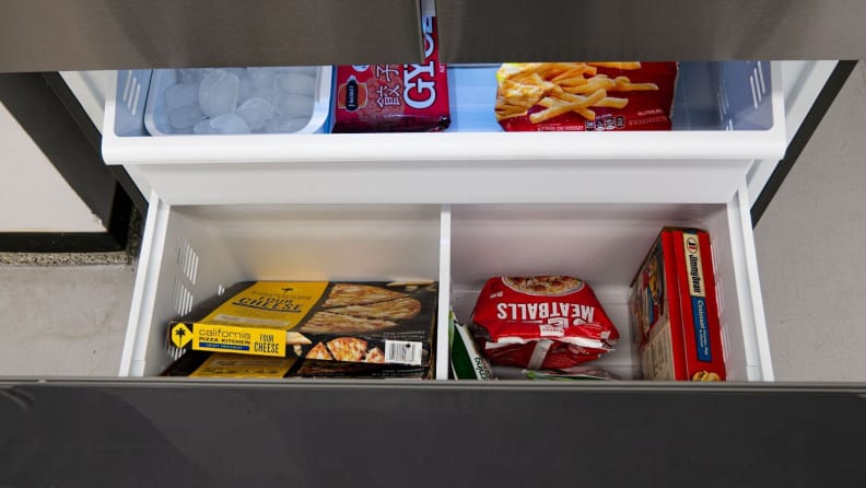 A close-up of the fridge's open freezer drawer, showcasing its main bin and the shelf that sits above it. The left corner of the upper shelf houses an ice reservoir. The rest of the space is filled with frozen foods.