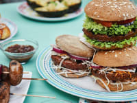 A spread with vegan burgers, skewers, and other out-of-focus plates with food on a blue tablecloth