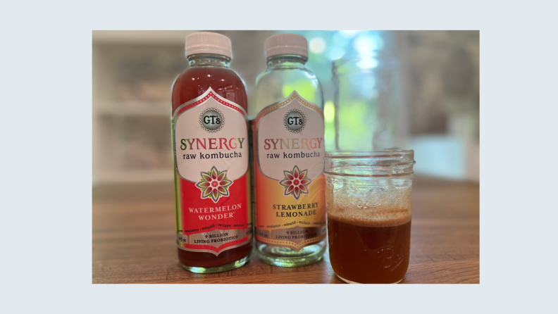 Two bottles of Synergy kombucha on a wood table with a small mason jar of kombucha.