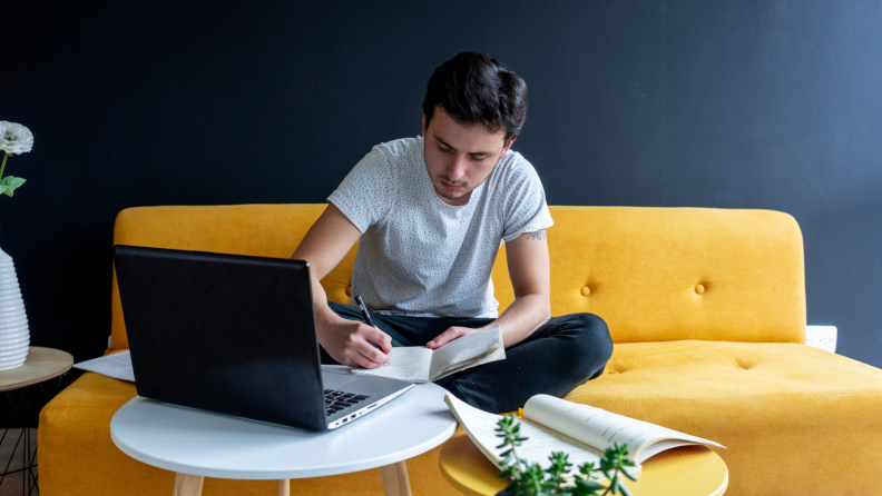 A young writer, or student, sits working on a yellow couch. There’s a laptop on the table in front of them; their focus is on a small notebook.