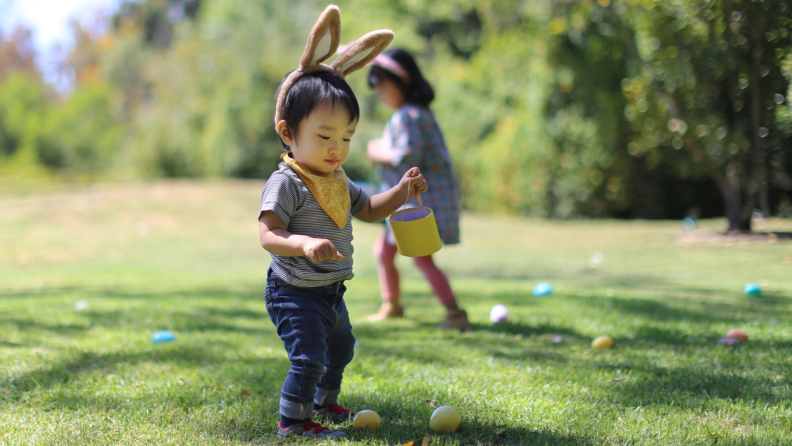 A young child wearing fake rabbit ears picks up Easter eggs on a grass field.