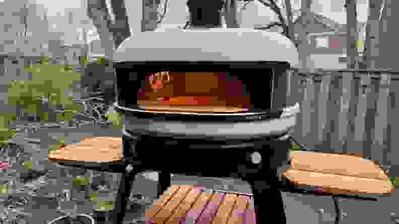 Gozney Dome Pizza Oven on stand with wood side tables, outdoors with open flame inside.