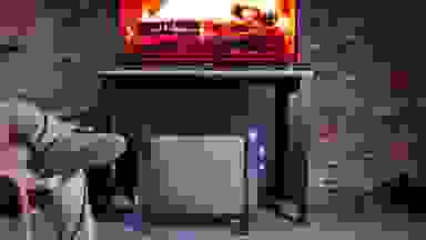 A space heater in a living room next to a person in a living room with a TV displaying an image of a fireplace
