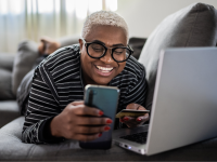 A person of color lays on a couch with a laptop and smartphone